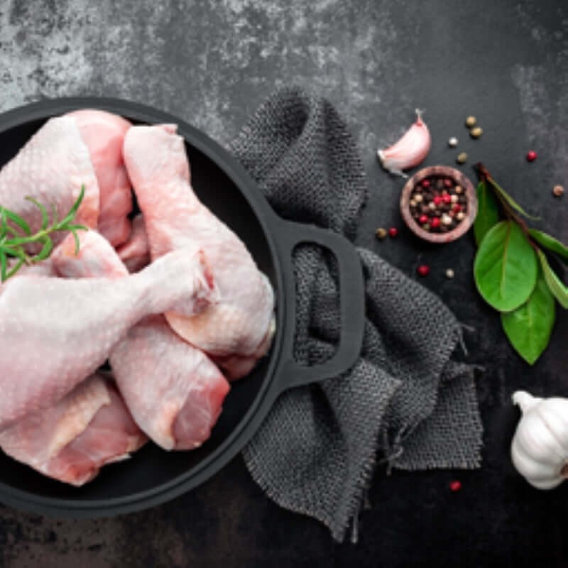 Premium chicken drumsticks in a black pan with herbs and spices on a textured surface.
